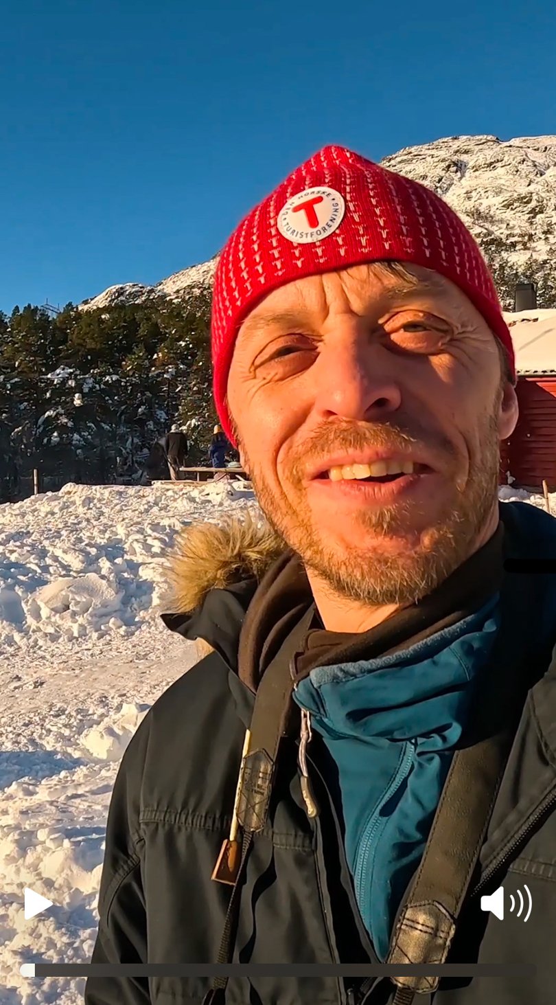 Person smiler til kamera foran rød hytte.