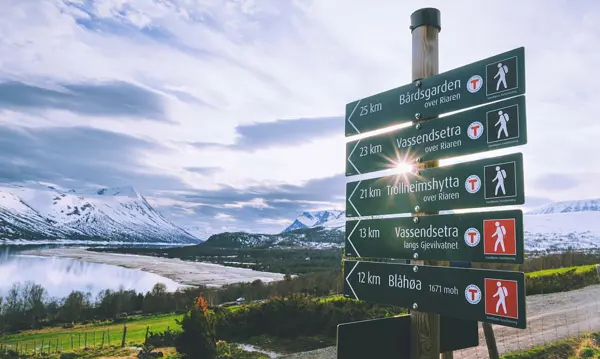 Bildet viser en skilt-stav, der fem forskjellige skilt peker veien og avstanden til andre lokasjoner. Fra toppen: Bårdsgaden, Vassendsetra (over Riaren), Trollheimshytta, Vassendsetra (langs Gjevilvatnet), og Blåhøa. Delvis snødekket fjell i bakgrunnen, og mellom oss og der er det en innsjø. Solen skinner mellom to av skiltene.