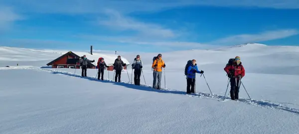 Ei hytte er nesten helt nedsnødd, man ser bare taket. Foran står det en gjeng skiløpere på rekke. De smiler. Det er fint vær og blå himmel.
