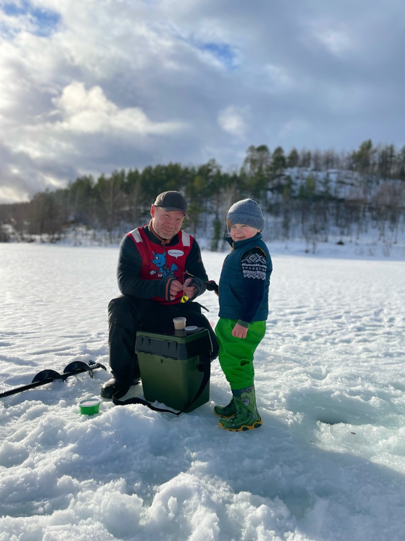 En mann sitter på en stol på isen og et lite barn står ved siden av. De har borret et hull i isen og har isfiskeutstyr. 