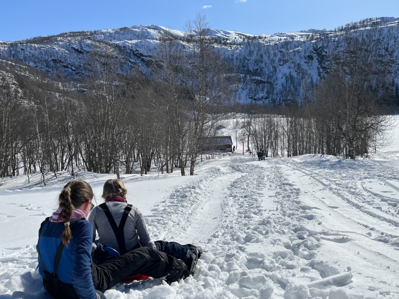 To jenter sitter på hvert sitt akebrett og skal til å ake ned en bakke. Det er trær lengre ned, et naust og et islagt vann.