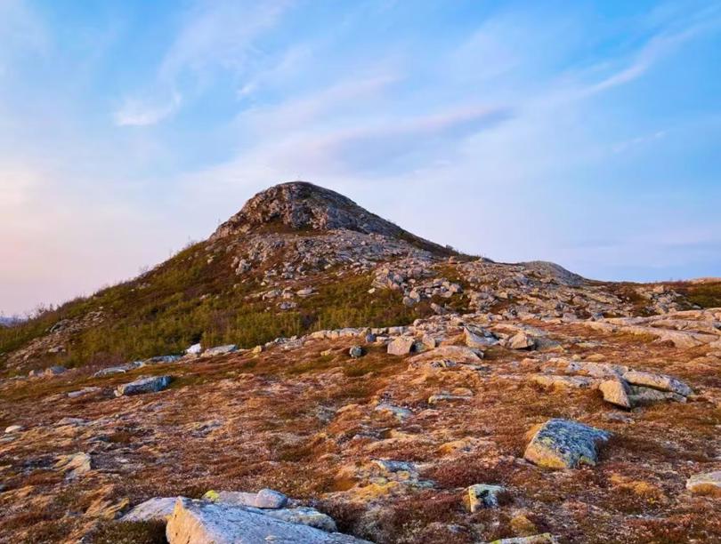 Bilde av en fjelltopp som opptar meste av bildet. Venste side av fjellsiden er dekket av grønn vegetasjon. Sommer eller vår. Skyfri himmel. En slette med litt gul vekst og store steiner strødd rundt er i forgrunnen. 