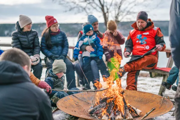 Barn og voksne sitter ute på vinteren rundt bålet og koser seg og griller pølser.