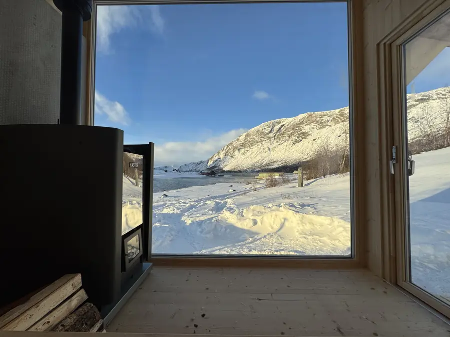En koselig vinterutsikt fra innsiden av en hytte med store vinduer, hvor snødekte fjell og et rolig vann preger landskapet. Vedovnen står klar med vedstabler, og gir en følelse av varme i det kalde vinterlandskapet.