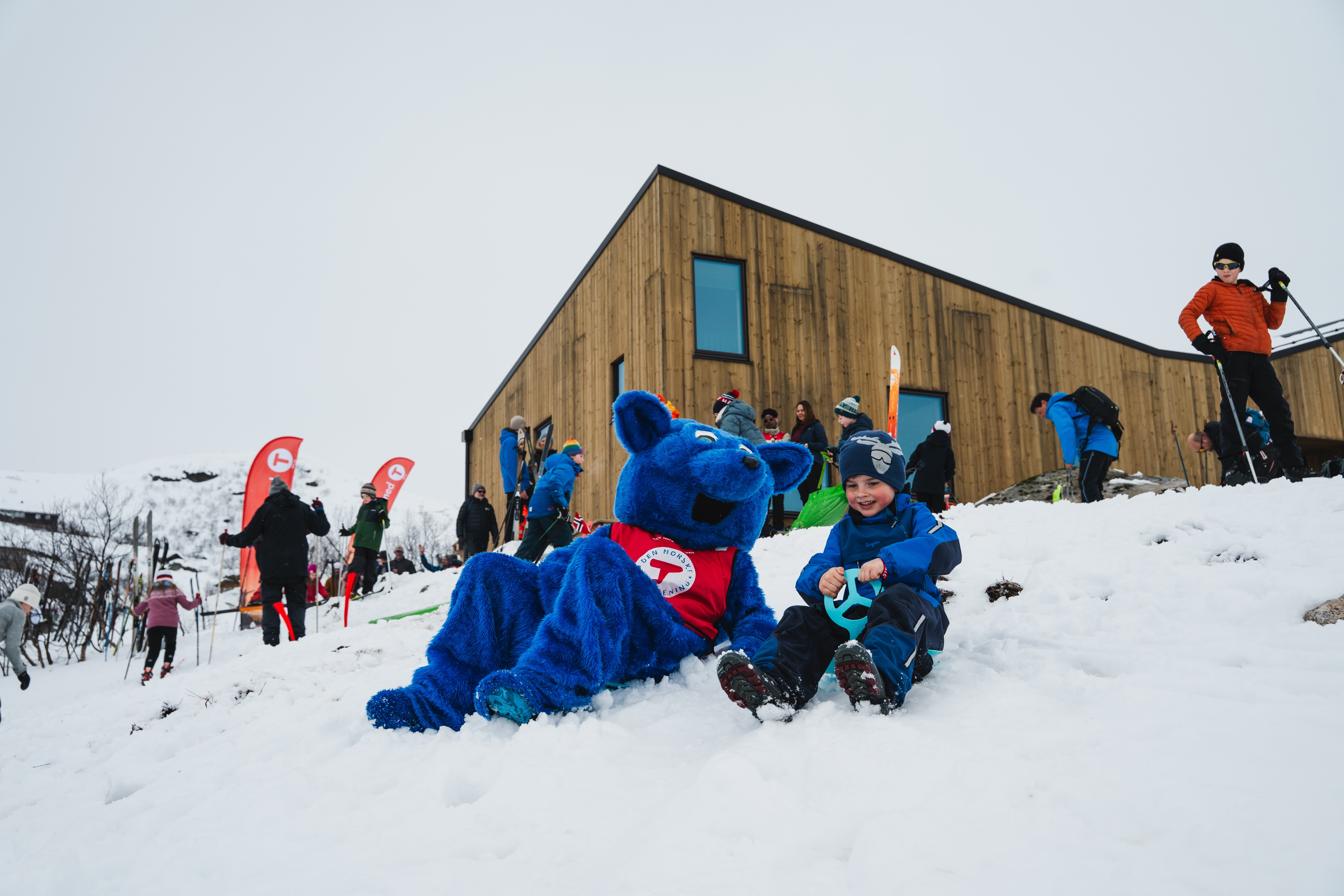En gutt i bl&aring; dress, og den bl&aring; fjellreven Turbo, Barnas turlag sin maskot, aker i sn&oslash;en utenfor Turbohytta. Noen folk i bakgrunnen foran en ny og moderne hytte. 