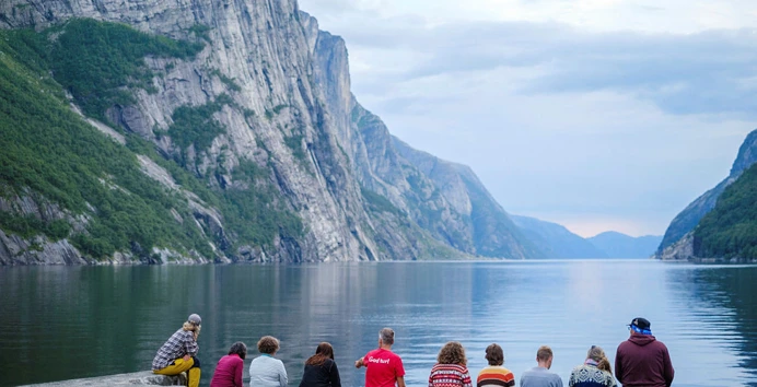En gruppe mennesker sitter med ryggen til på brygga innerst i en lang fjord.