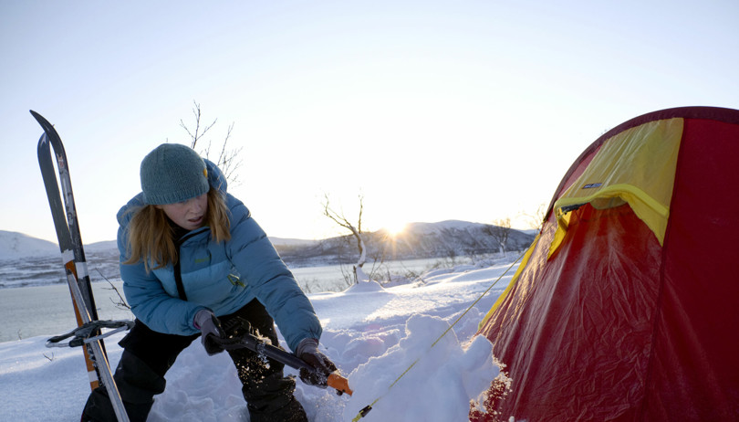 Person med blå dunjakke spar snø over kantene på et rødt og gult telt.