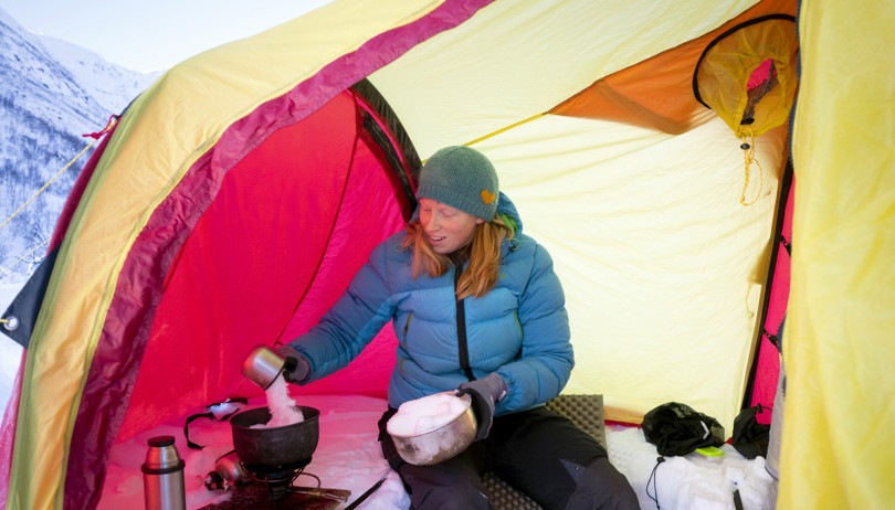 Person i blå dunjakke sitter i forteltet på et rødt og gult telt og heller snø opp i en kjele på et stormkjøkken.