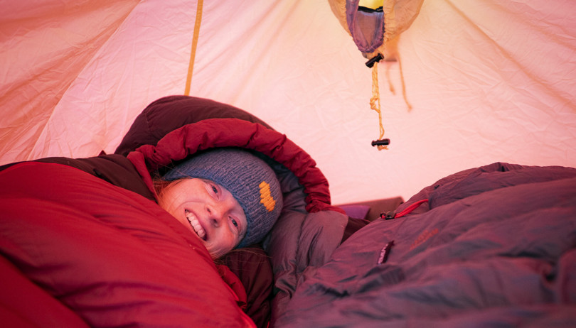 Person med blå lue ligger i en rød sovepose i et rødt telt. 