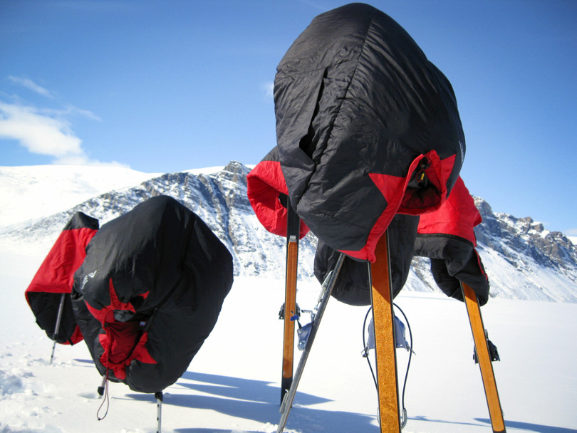 To svarte og røde soveposer henger over ski og staver som står oppreist i snøen.