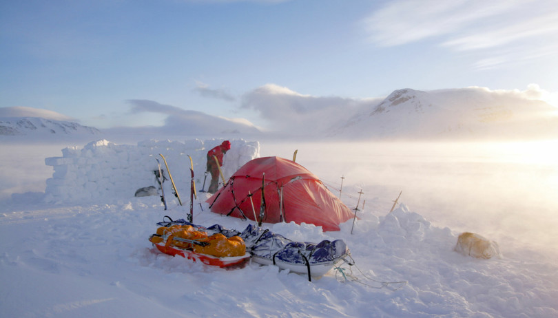 Rødt telt står inntil en levegg av snø på fjellet. Det føyker snø rundt. Bak teltet står en person i rød jakke. Foran tiltet ligger to pulker i snøen.