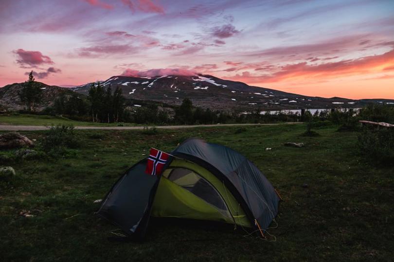 Et telt er oppslått på en gresslette, og i bakgrunnen ruver et stort fjell under en rosa himmel.