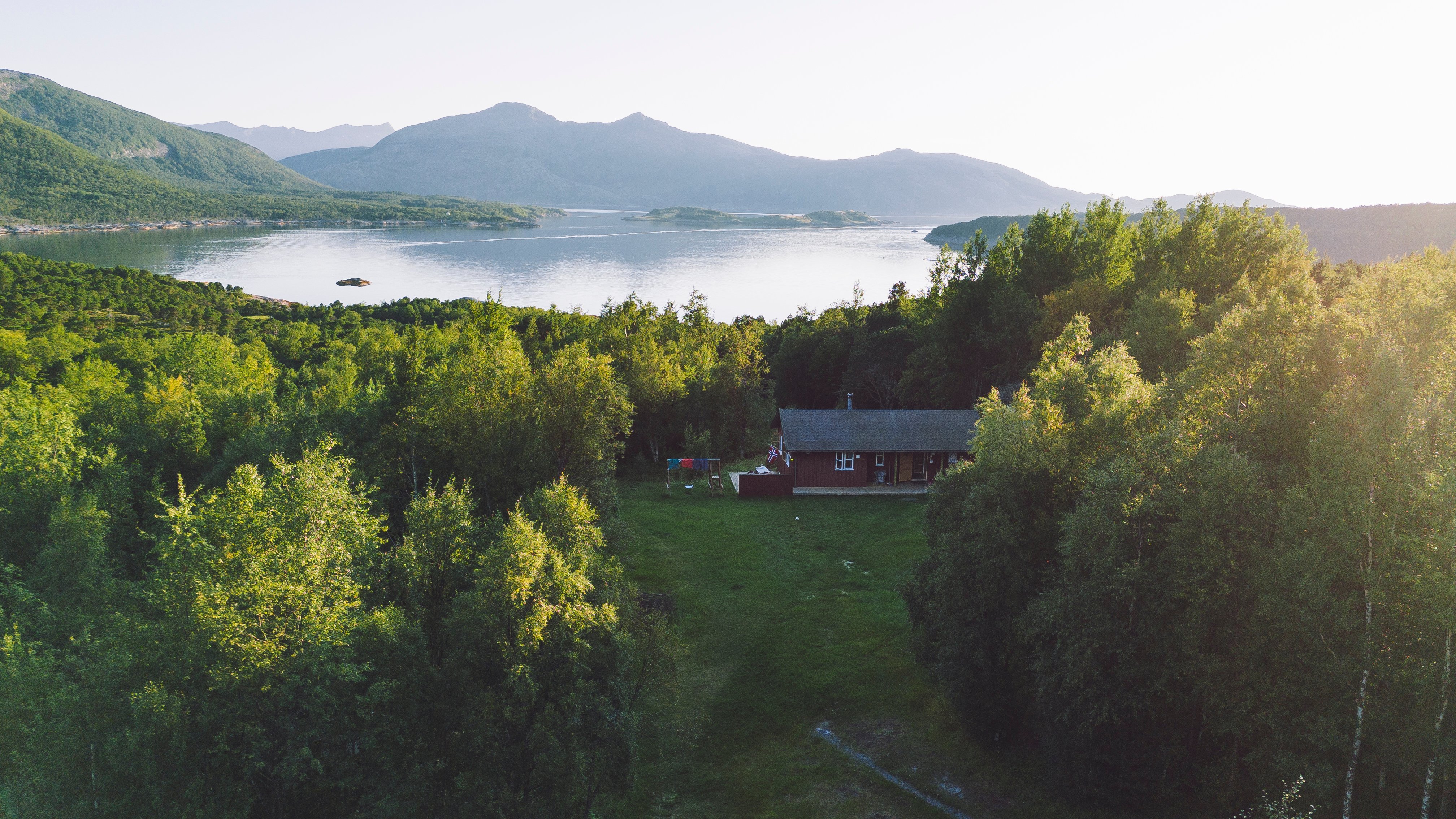 Et kystområde med hav i bakgrunnen, skoger foran og ei hytte liggende i skogen med en gressplen foran.