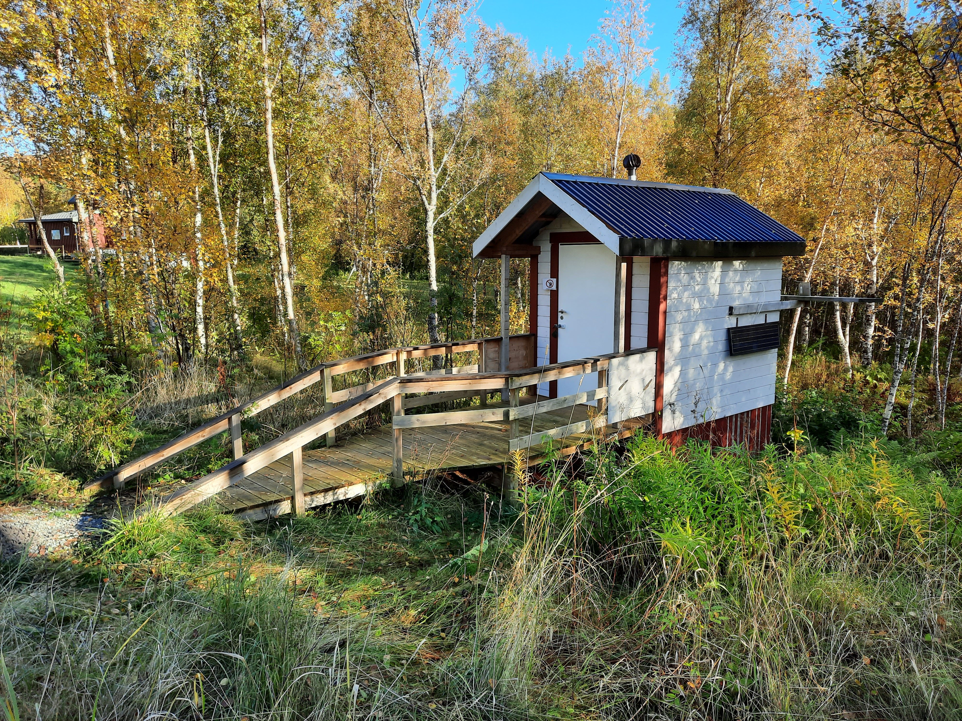 Ei hvitmalt utedo med trefarget ramp foran. Det er gress og trær med høstfarger rundt.