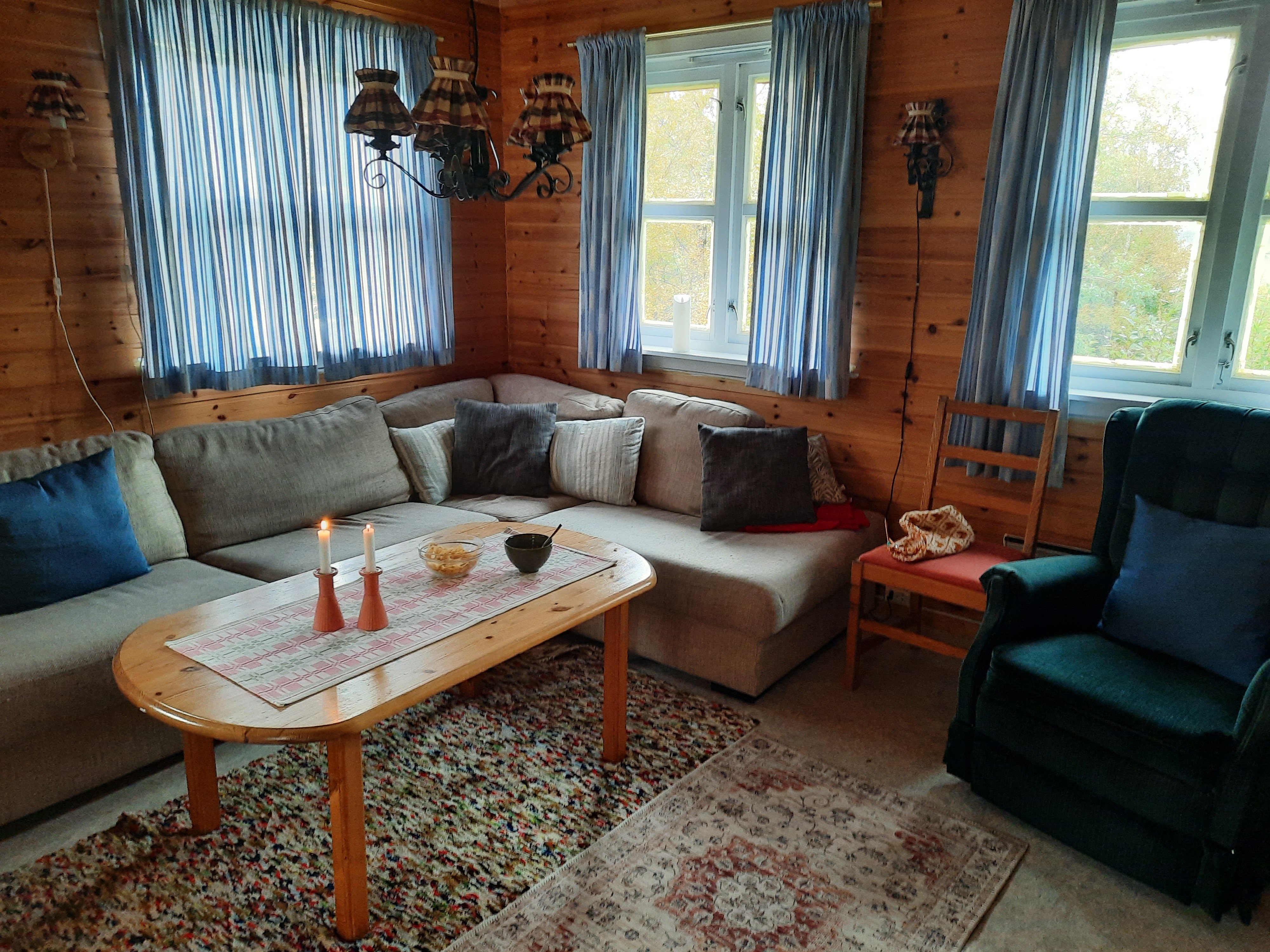 Ei hyttestue med trehvite vegger, en stor hvit hjørnesofa med bord foran og en mørk lenestol.