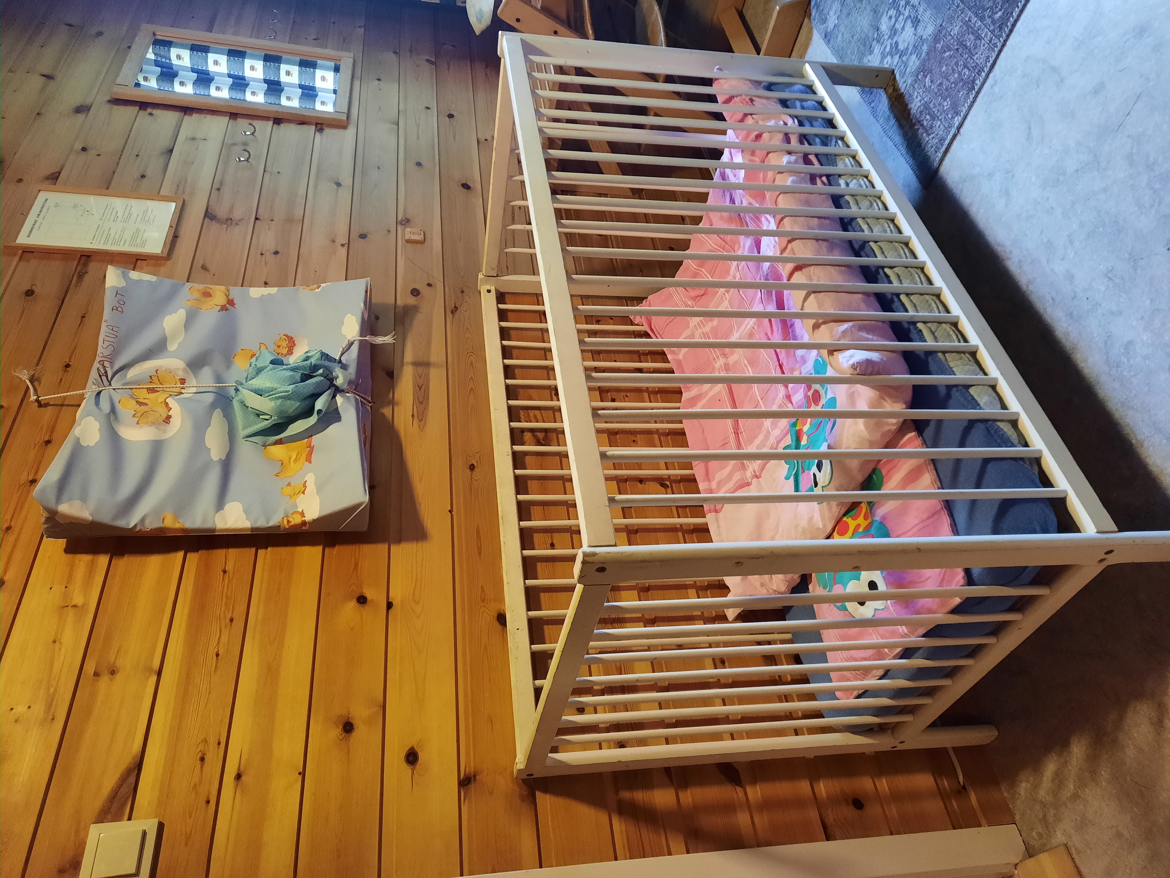 Ei hvit sprinkelseng med rosa pute og dyne står foran en trehvit vegg.