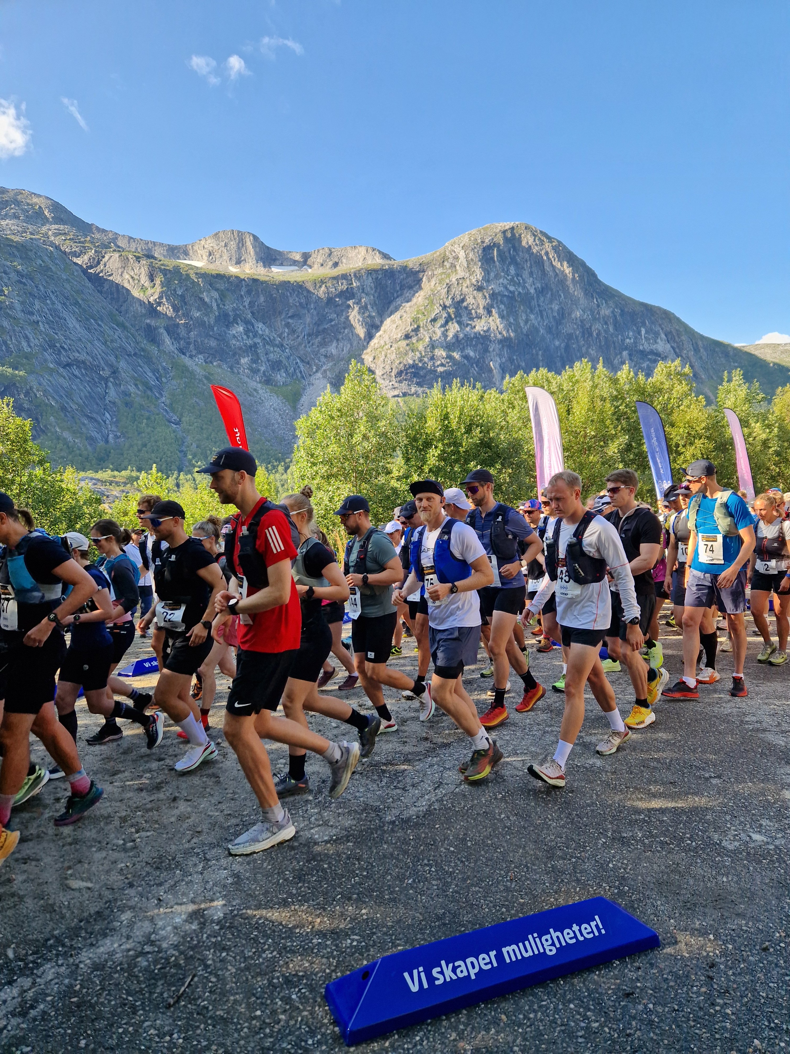 En gjeng folk med løpeklær i ulike farger løper. Det er trær og fjell i bakgrunnen. 