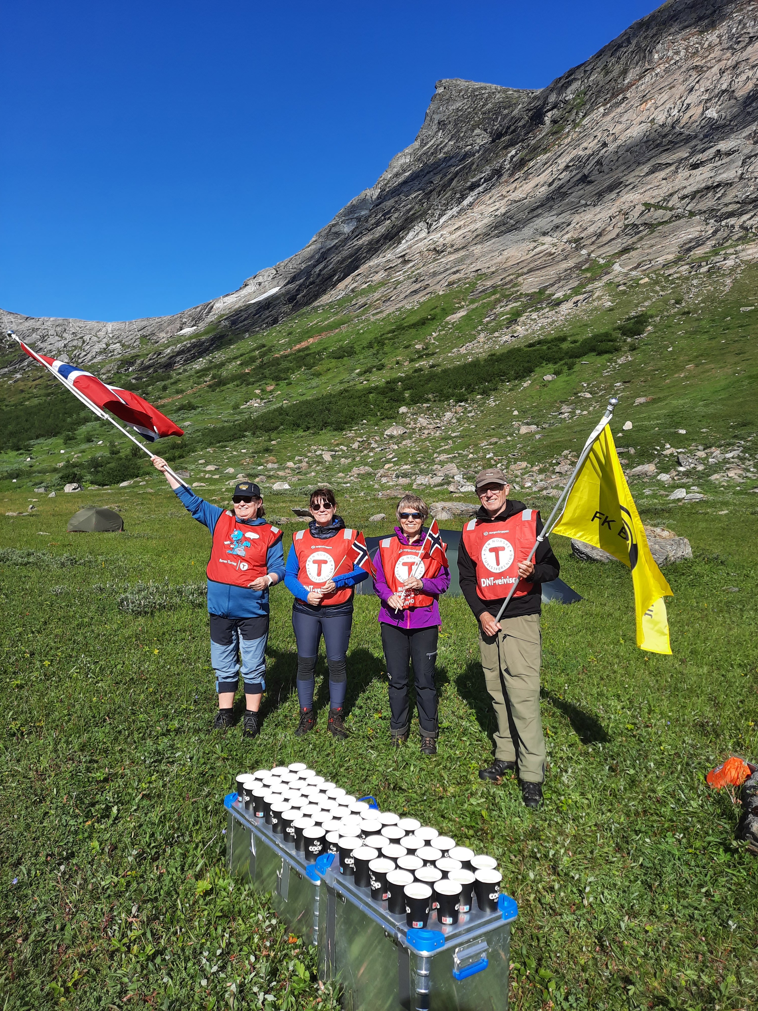 Fire mennesker med røde vester, tre damer og en mann, står på en gresslette. En av dem holder et norsk flagg og en et gult flagg. Det er grå fjell i bakgrunnen. Foran dem står en kasse med mange drikkebegre på.