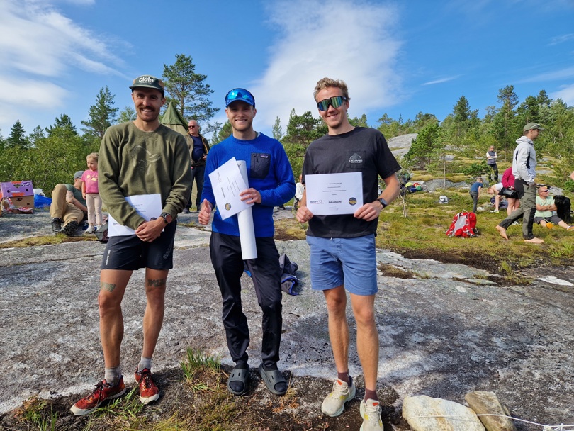 Tre menn står ved siden av hverandre og smiler. En av dem har grønn genser og caps, en har blå genser og svart bukse, og den siste har svart t-shorte og blå shorts. De holder et ark i hendene. Bak er det noen mennesker og trær.