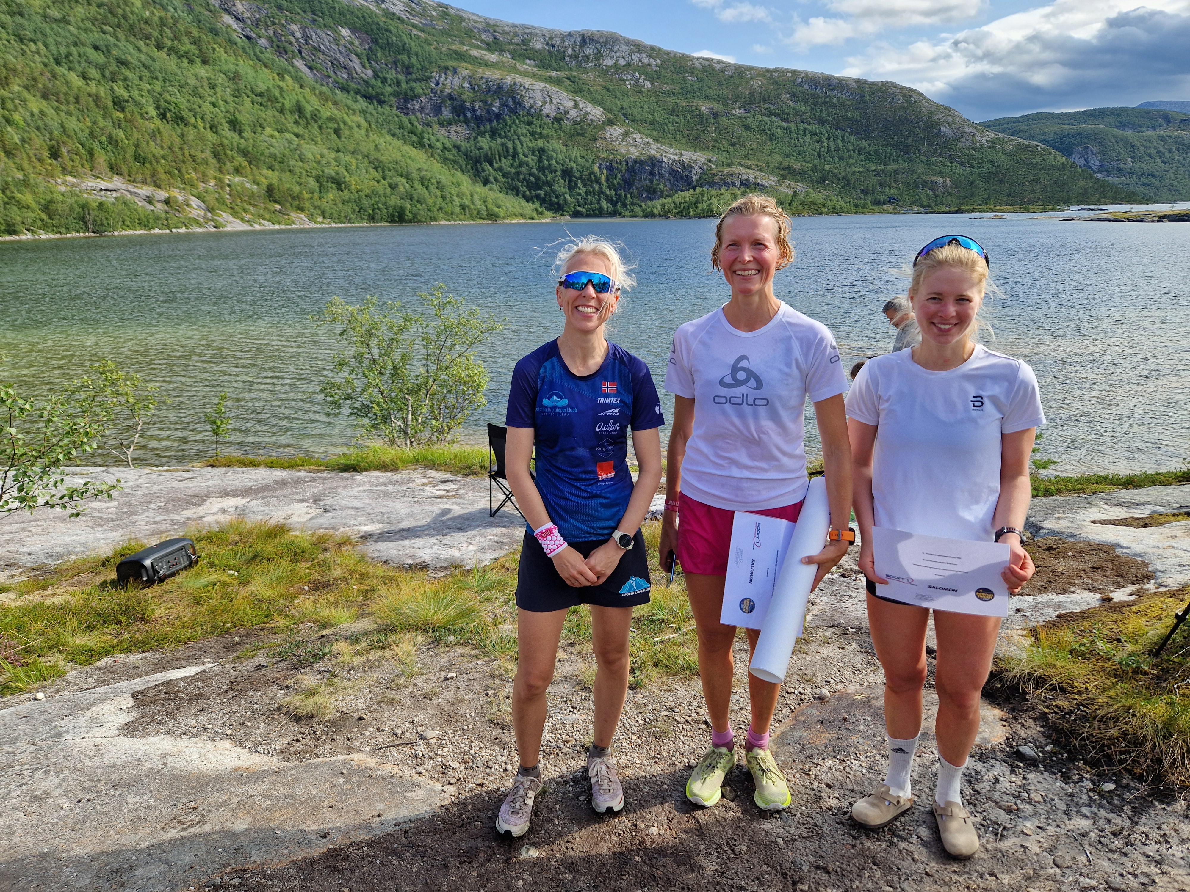 En dame i blå t-skjorte står ved siden av to damer i hvite t-skjorter. De har løpeshorts og løpesko. Alle tre smiler. Bak dem er det et vann, skog og fjell. 