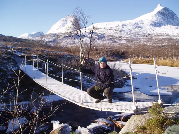 Bru med en person på og snøkledte fjelltopper i bakgrunnen