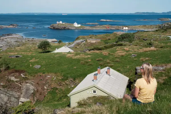 En kvinne sitter i gresset og ser ut over havet. En lysegrønn hytte er nedenfor.