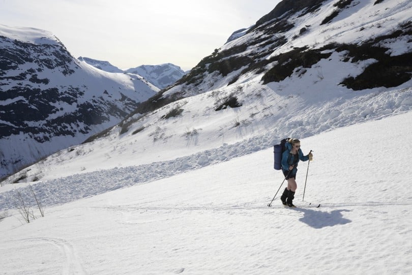 Person i shorts og med stor sekk på ryggen går opp ei bratt fjellside på ski. 