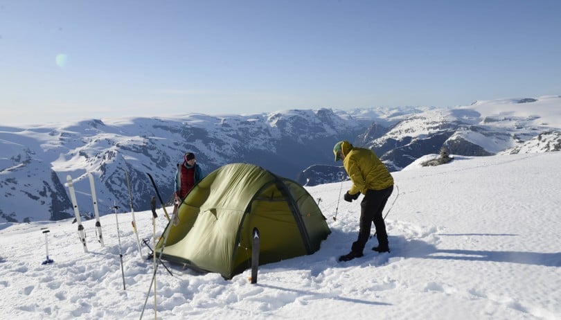 To personer setter opp et grønt telt i snøen høyt til fjells.