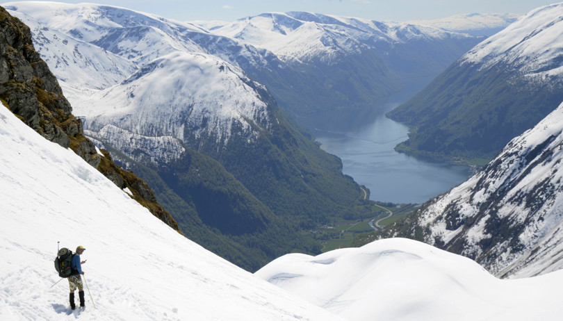 Person på på ski i en bratt fjellside skuer utover en fjord omkranset av fjell. Fjellene er snøkledde, nede i dalen og ved fjorden er det bart og grønt.