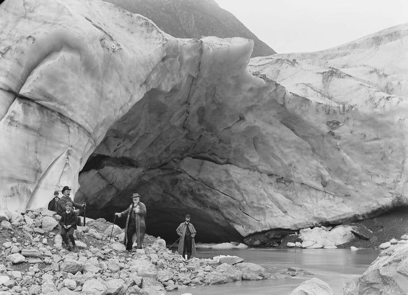 Gammelt bilde som viser fem menn stående ved elvemunningen under en høy isbrekant.