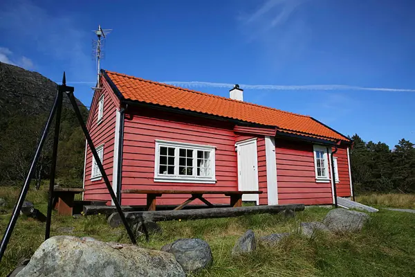 Rød hytte med bålpanne og benk foran, blå himmel bak.