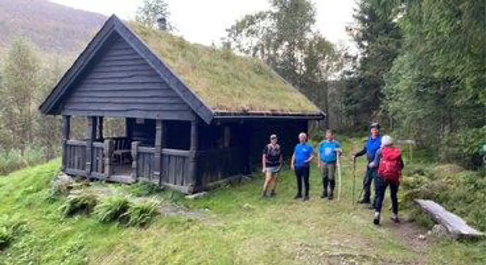 Fire turgåare ved ei brunbeisa enkel hytte med torv på taket 