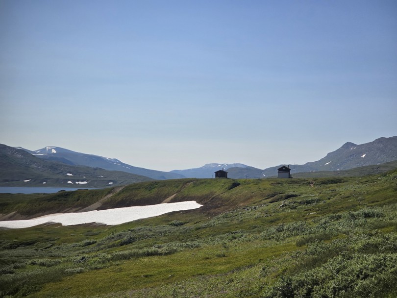 Et høyfjellslandskap med lyng og en diger snøflekk. Fjell på begge sider. To rødbrune hytter ligger i bakgrunnen.