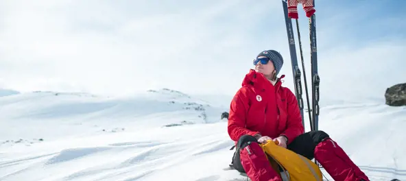 Kvinne i rød jakke sitter i snøen. Hun ser fornøyd ut. Ved siden av henne ses ski og en oransje tur sekk