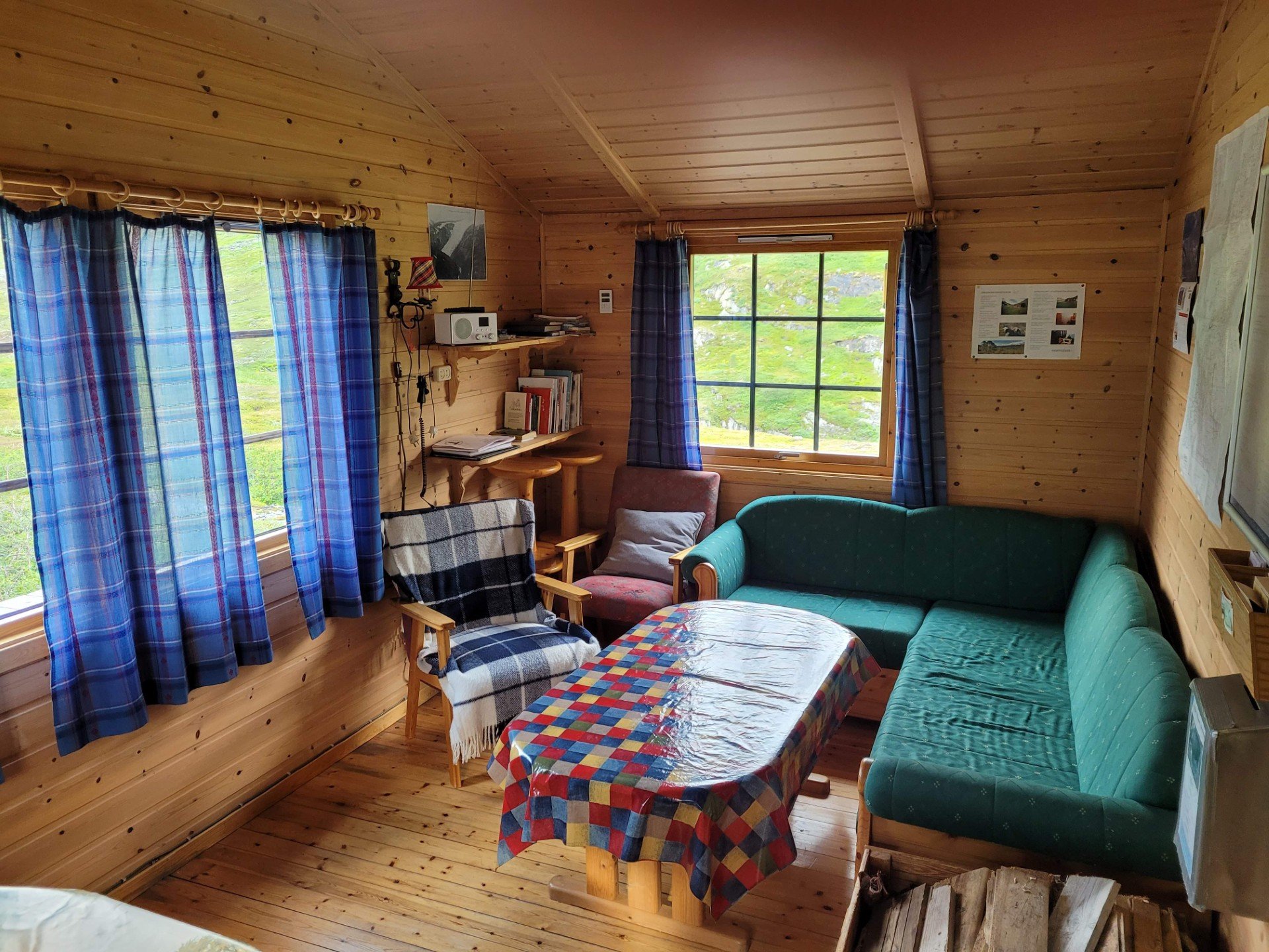 Ei hyttestue med grønn hjørnesofa, bord med fargerik duk, to lenestoler og ei vedkasse.