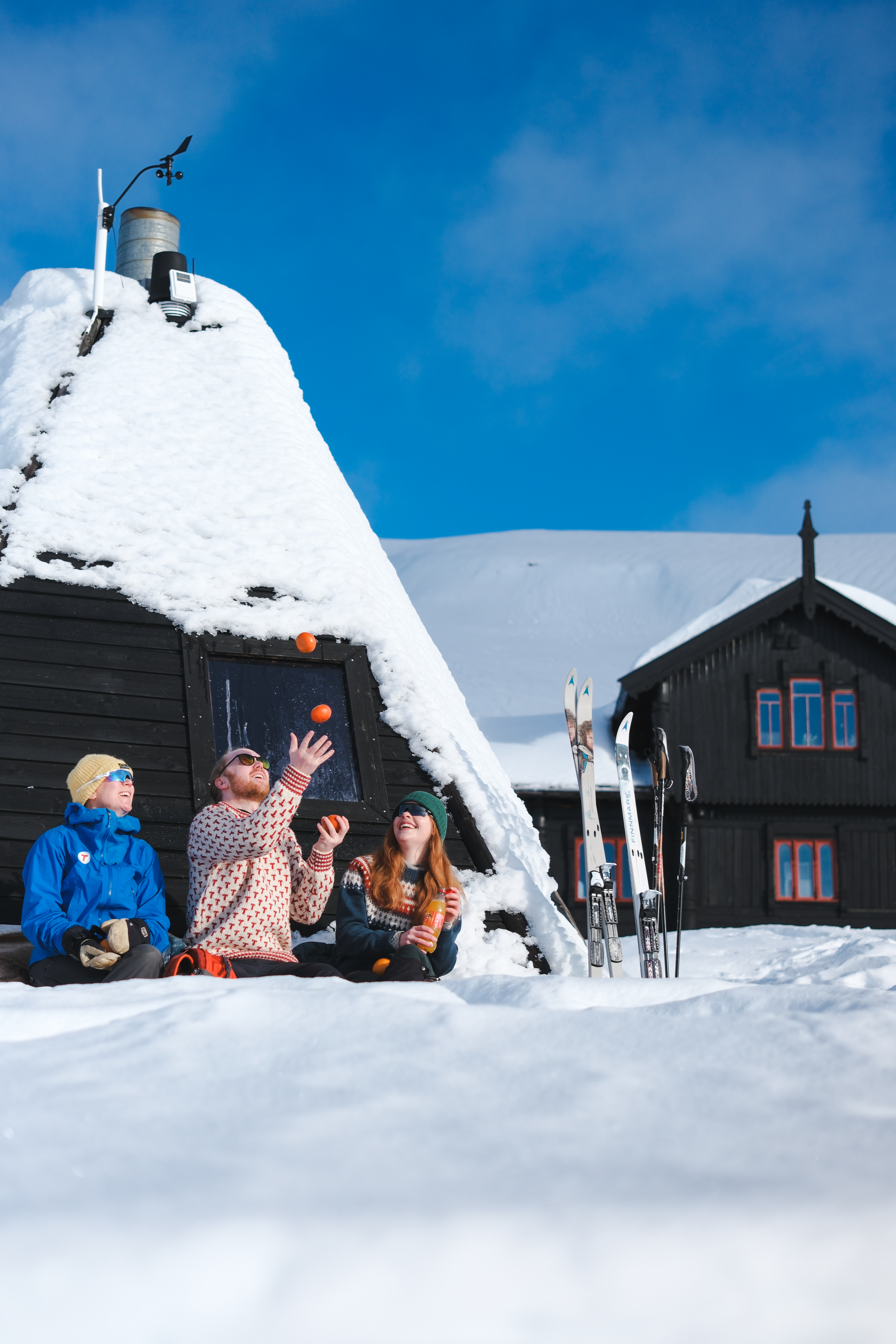 Tre personer med ullgenser og appelsinger sitter i en solvegg, sn&oslash; og bl&aring; himmel