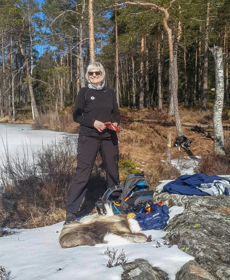 En dame ved et vann i skogen, med reinsdyrskinn og sekk på bakken.