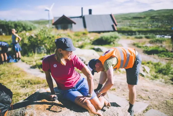 En mann med oransje refleksvest ser på hælen til en kvinne som sitter på en stein.