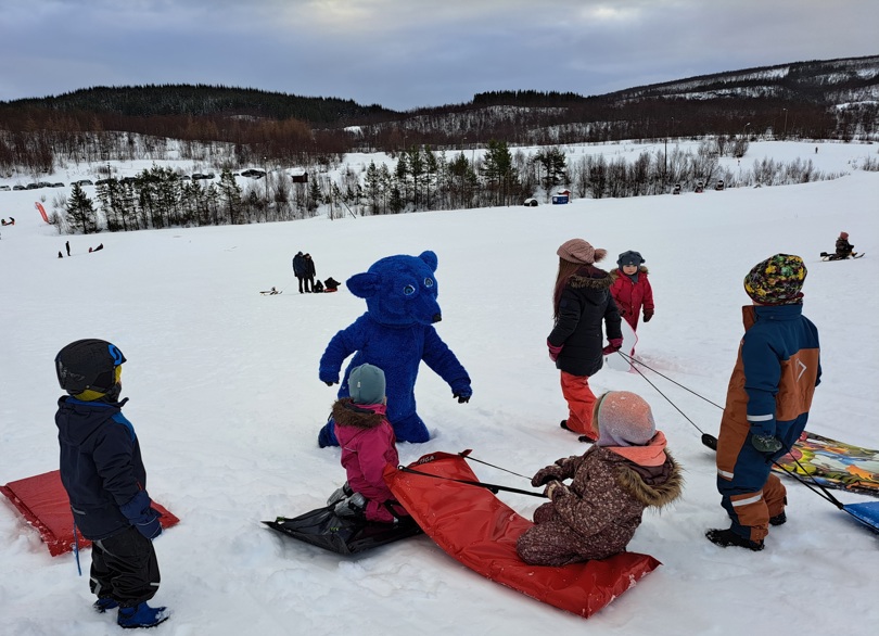 En stor, blå rev leker med små barn på akebrett. Det er vinter og snø, trær og folk i bakgrunnen.