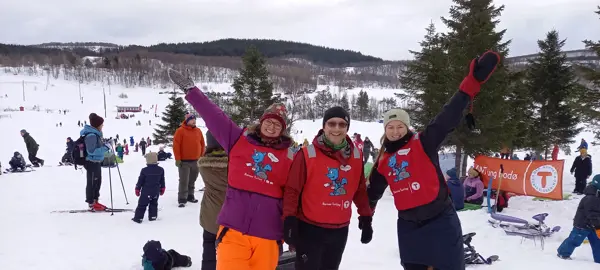 Tre personer med rød vest har en hånd i været. De ser veldig glade ut. Det er vinter og snø på bildet. Folk og trær i bakgrunnen. 