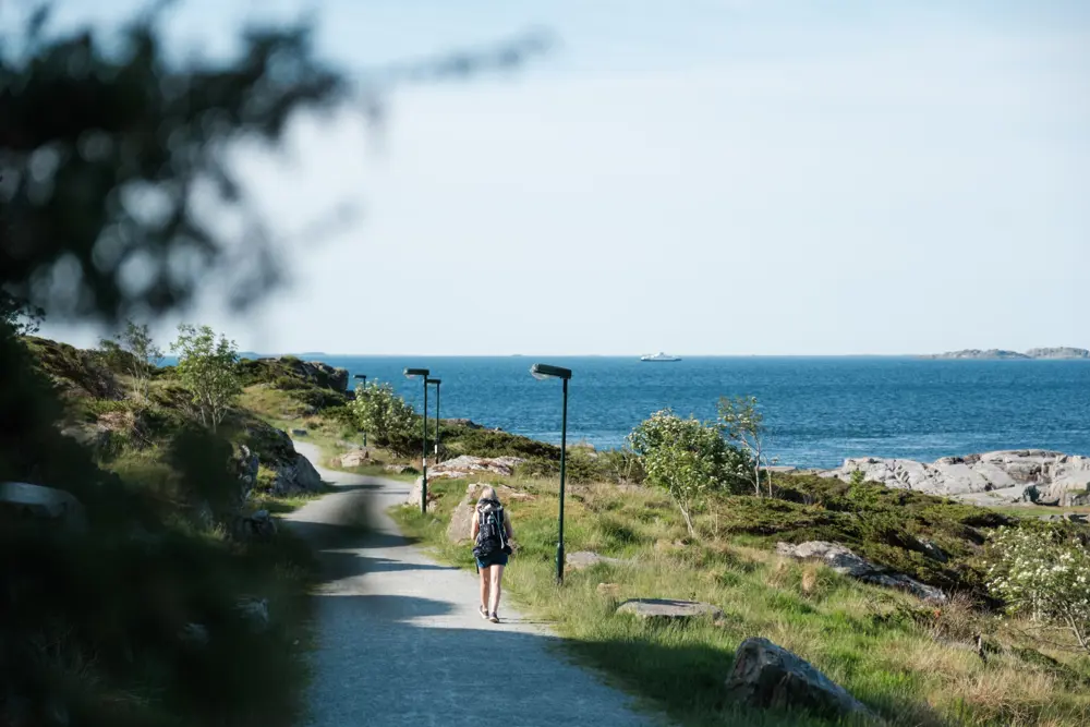 En kvinne med ryggsekk går innover en grusvei, med havet på høyre side.