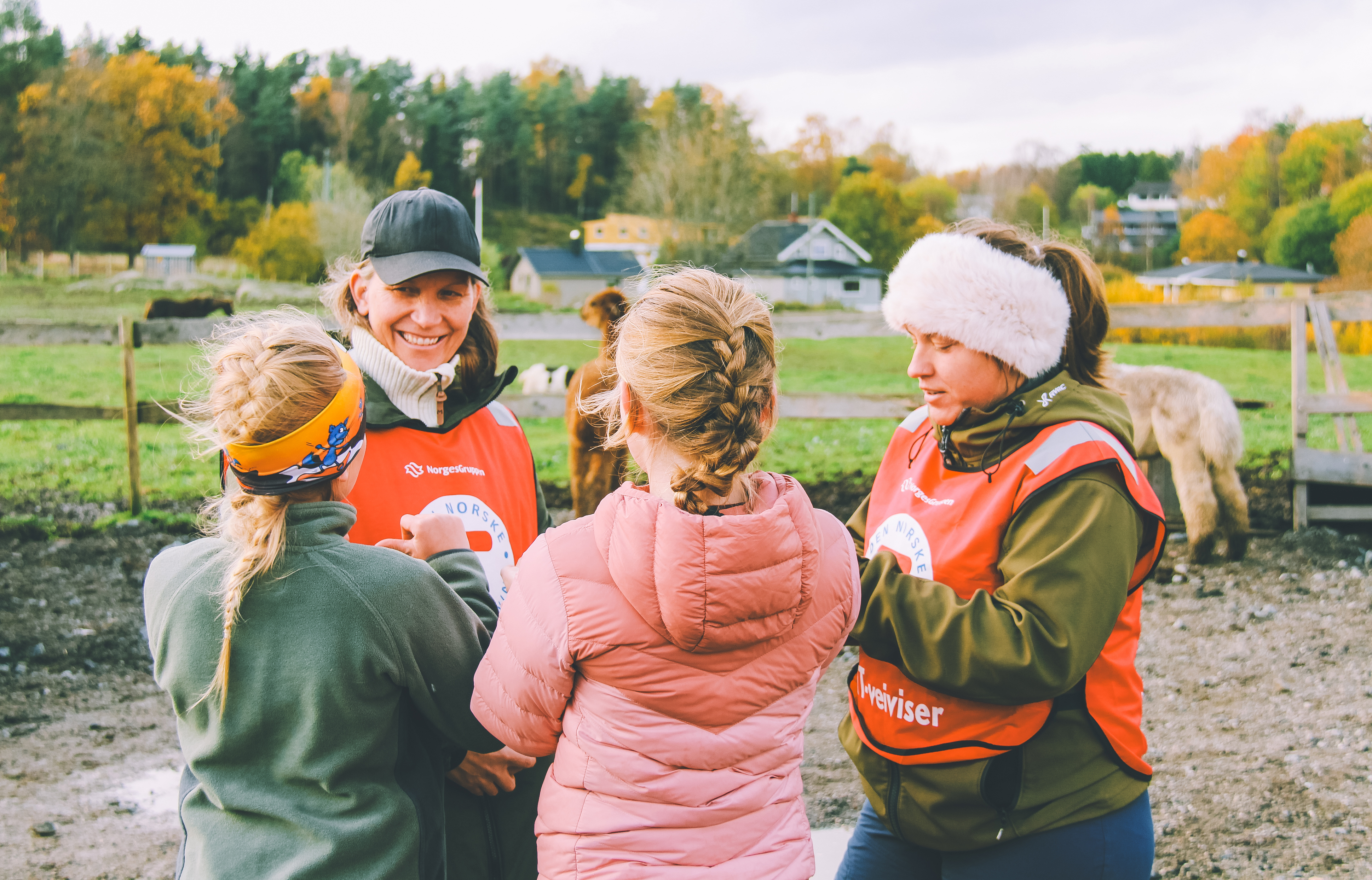 Fire personer iført friluftsklær og røde vester står sammen og snakker på et gjørmete jorde, med trær og hester i bakgrunnen.