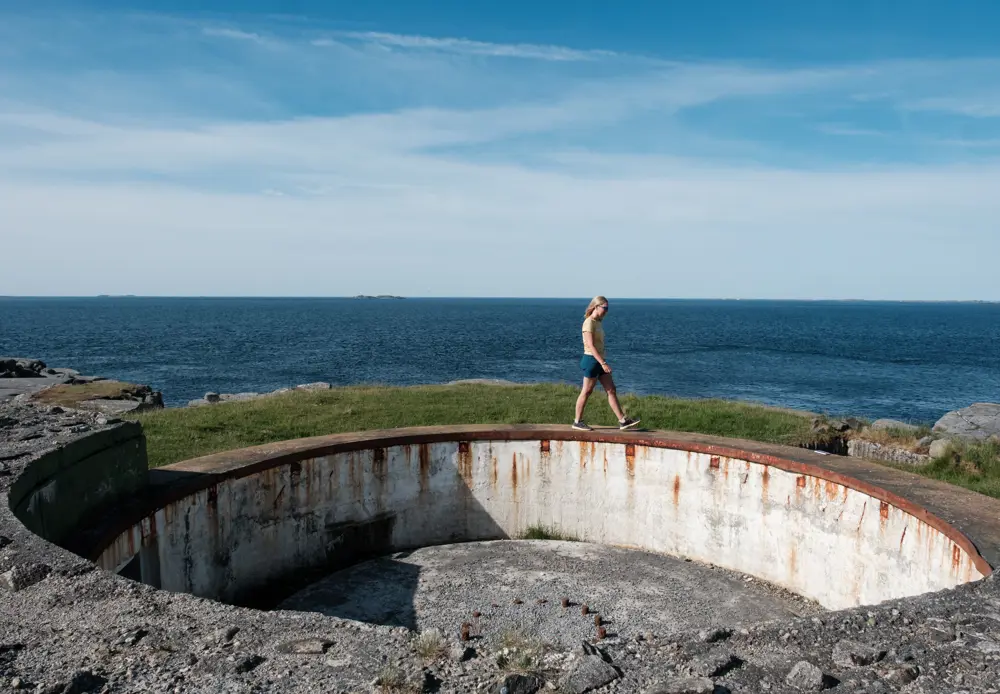 En kvinne i gul tskjorte og shorts går på toppen av en bunkers. Bak henne skimtes blått hav.