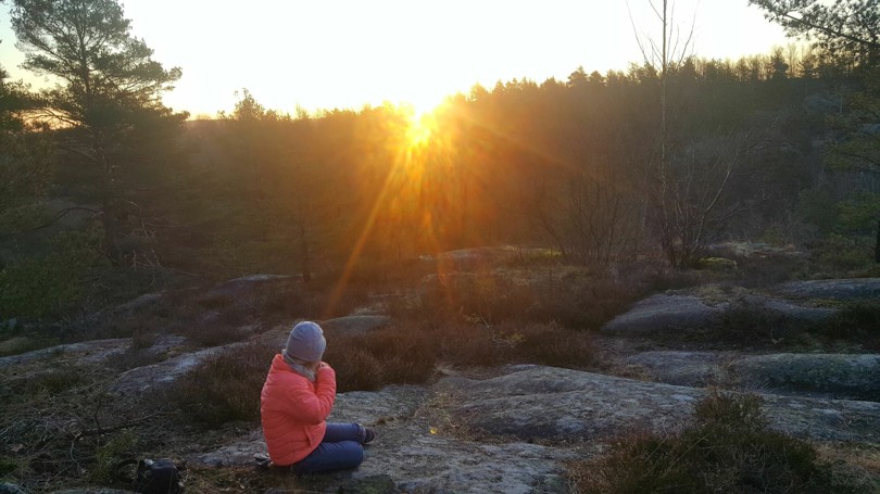 Person sitter på et svaberg i skogen og ser mot sola som stiger opp over skogen i horisonten.