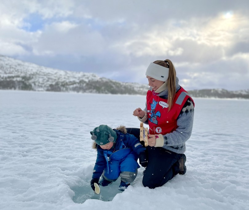 Ei jente og en liten gutt isfisker. Det er snø og is på bildet. Jenta har på seg en rød vest, og gutten har på seg en blå dress.