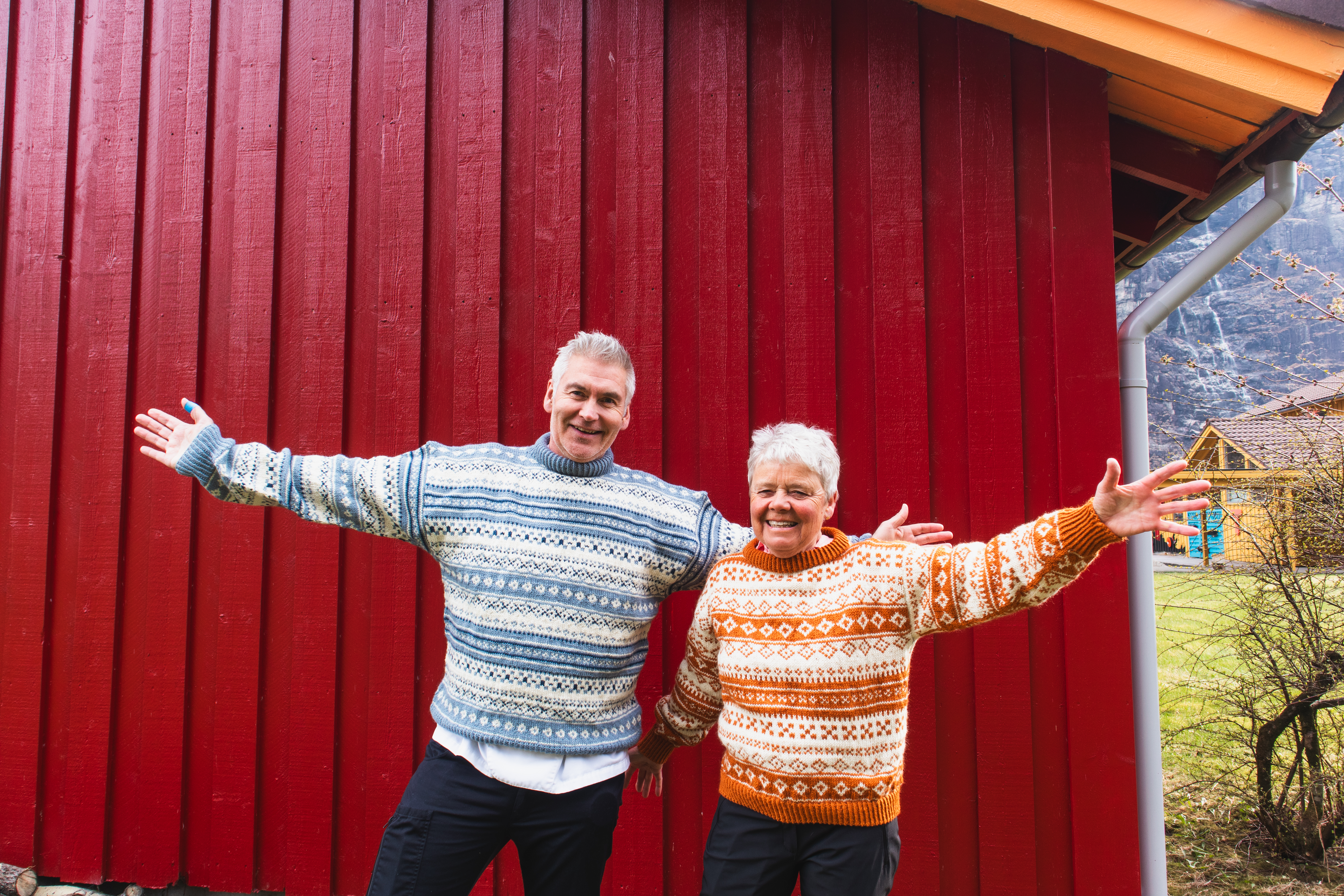 En mann og en dame med m&oslash;nstrede strikkegensere foran en r&oslash;d husvegg.