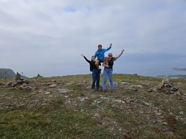 Tre ungdommer står på toppen av et fjell. En av dem sitter på skuldrene til de to andre. De holder armene i været og smiler. 
