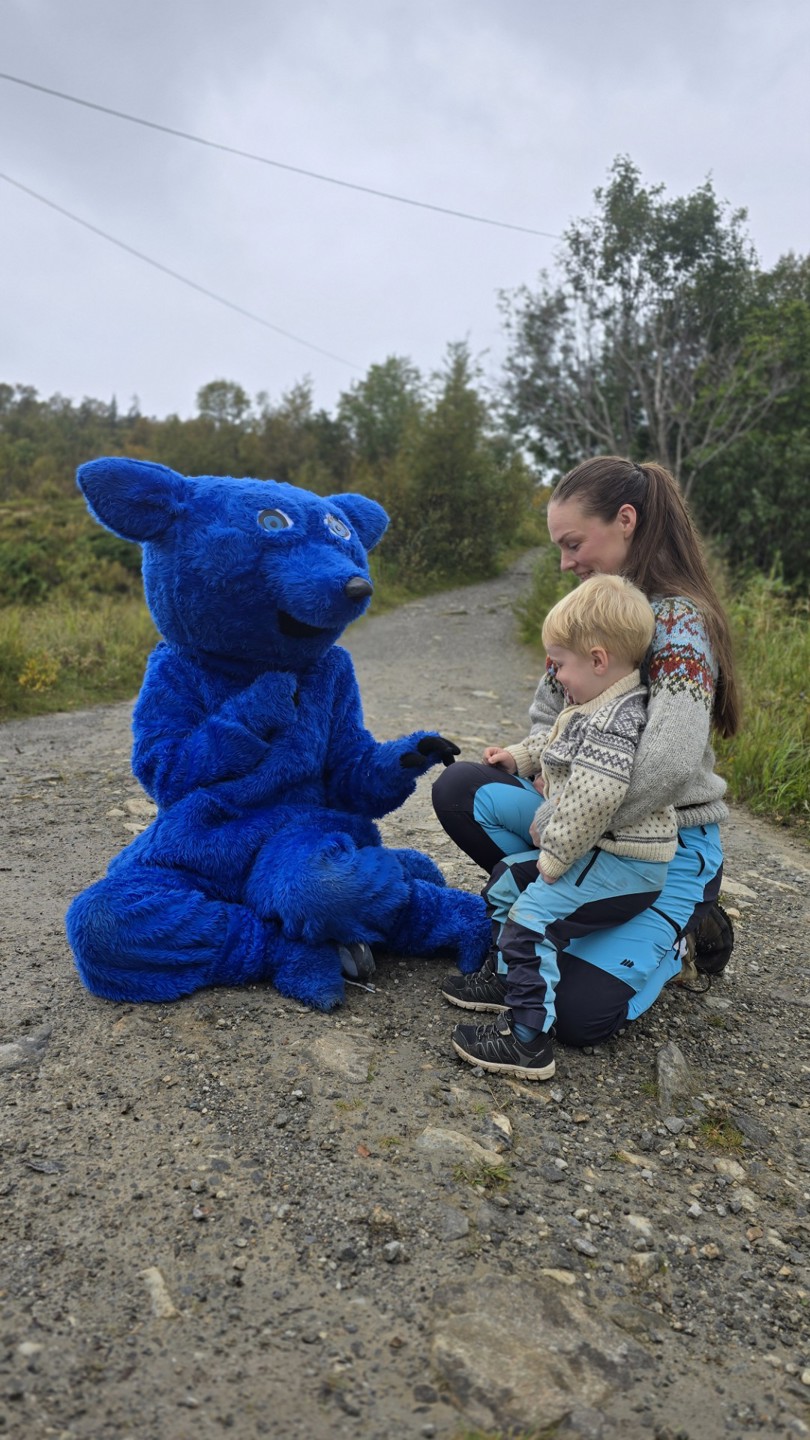 En dame med en liten gutt på fanget hilser på en stor blå bamse, som er Turistforeningens maskott.