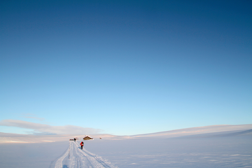 Skiløper på snødekt vidde - hytte i det fjerne.