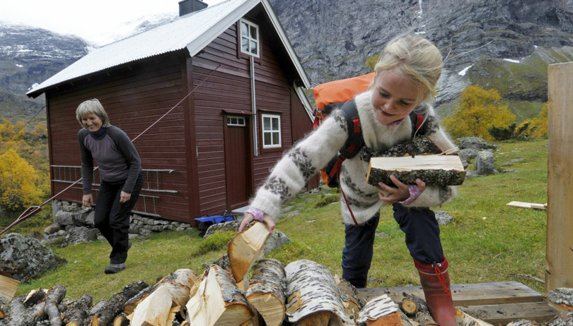 Jente med ullgenser og oransje sekk plukker opp vedkubber utenfor en brun hytte.  